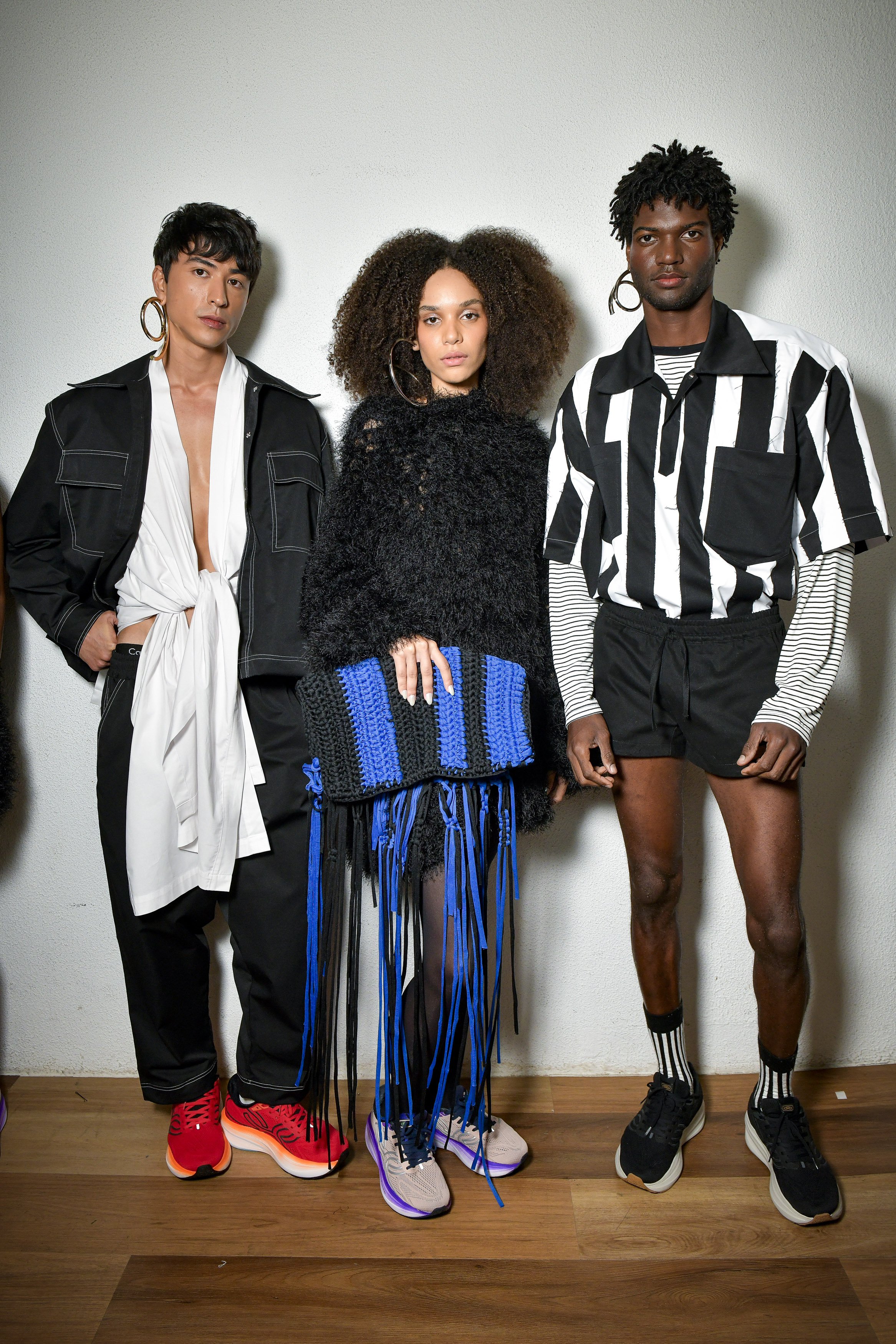 Led- SPFW N59Foto: Gustavo Scatena/ @agfotosite Imagem de três modelos no backstage da LED, na SPFW N59. Um modelo veste camisa branca aberta e desconstruída com blazer, a do meio veste vestido preto de pelos com bolsa de crochê de malha azul e o do canto direito, uma camisa de gola com listras verticais preto e branco e short preto.
