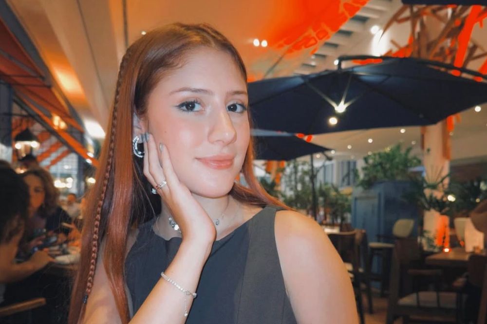 Imagem da influenciadora Liz Macedo, jovem branca e ruiva, com uma mão apoiada no rosto e usando um vestido preto sem manga.