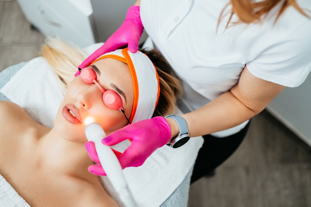 Foto de garota deitada em maca com proteção nos olhos e faixa no cabelo enquanto faz um tratamento de laser no rosto