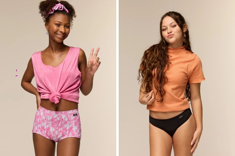 herself + ch Duas jovens posam em um estúdio com fundo neutro. À esquerda, uma garota usa um top rosa amarrado e uma calcinha estampada em tons de rosa, fazendo o sinal de paz e sorrindo. À direita, outra garota veste uma blusa laranja e uma calcinha preta, fazendo biquinho e gesto de paz com a mão. Ambas aparentam estar confortáveis e confiantes.