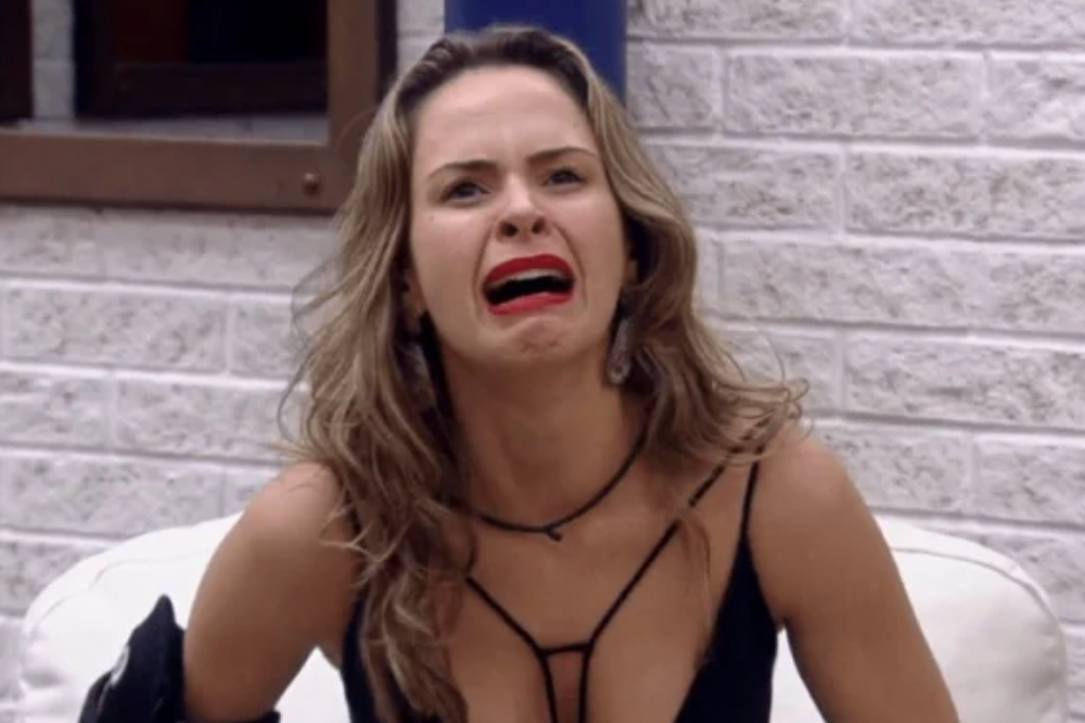 Mulher sentada em um sofá claro, usando blusa preta de alças finas e batom vermelho, aparece chorando com expressão de surpresa. Ela está em um ambiente interno com parede de tijolos brancos ao fundo e uma janela parcialmente visível.