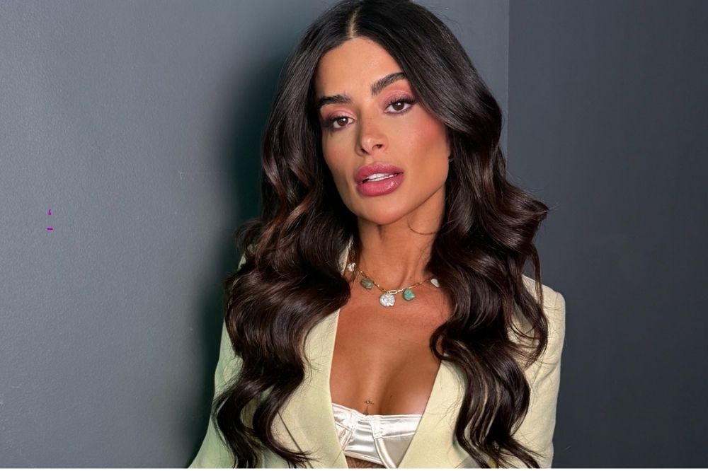 Mulher posa de frente para a câmera, com cabelos longos e ondulados castanhos, usando blazer claro sobre top estruturado e colar delicado, diante de uma parede cinza.