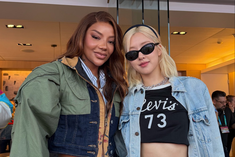 Duas mulheres posam juntas em um evento indoor: à esquerda, mulher negra com cabelo solto e jaqueta verde e jeans; à direita, mulher loira usando óculos escuros, top preto com a inscrição “Levi’s 73” e jaqueta jeans, ambas sorrindo para a câmera.