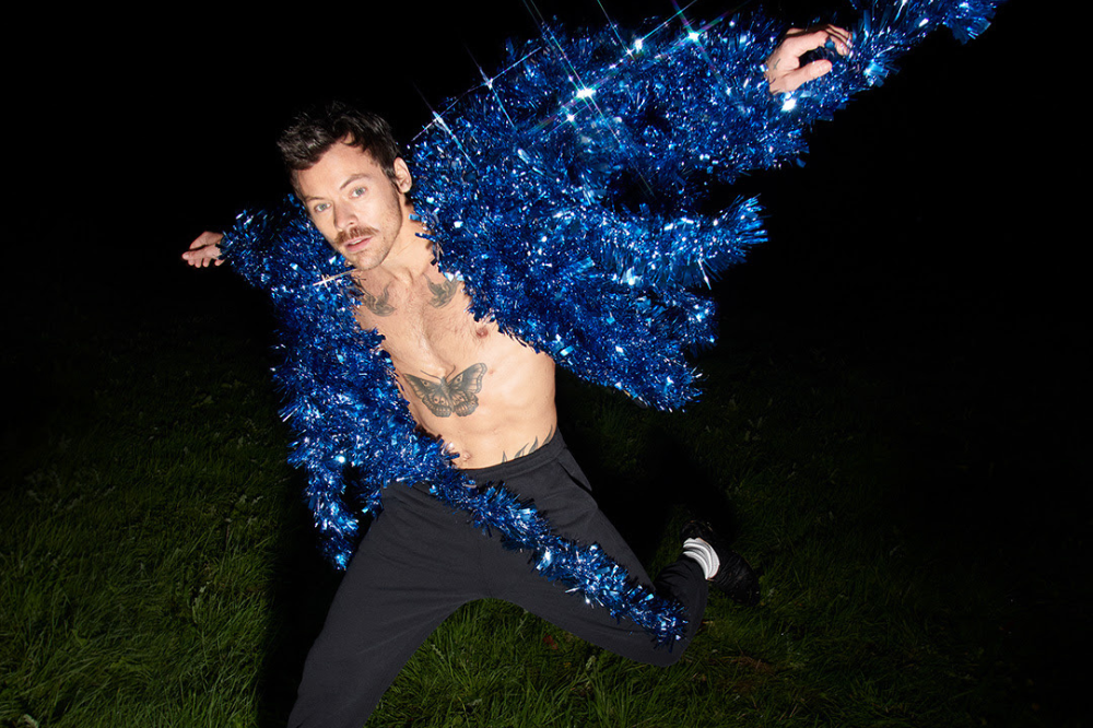 Homem salta sobre a grama à noite usando casaco azul brilhante de franjas, aberto no peito e exibindo tatuagens; ele está com os braços estendidos, em pose dinâmica, contra fundo escuro.