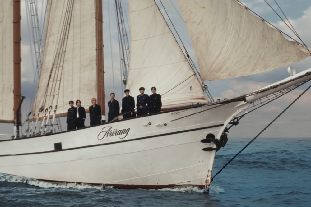 Sete jovens vestidos com roupas escuras e formais estão alinhados na proa de um veleiro branco chamado “Arirang”, navegando em mar aberto. As velas estão abertas e infladas pelo vento, enquanto o oceano azul e o céu parcialmente nublado formam um cenário calmo ao fundo.