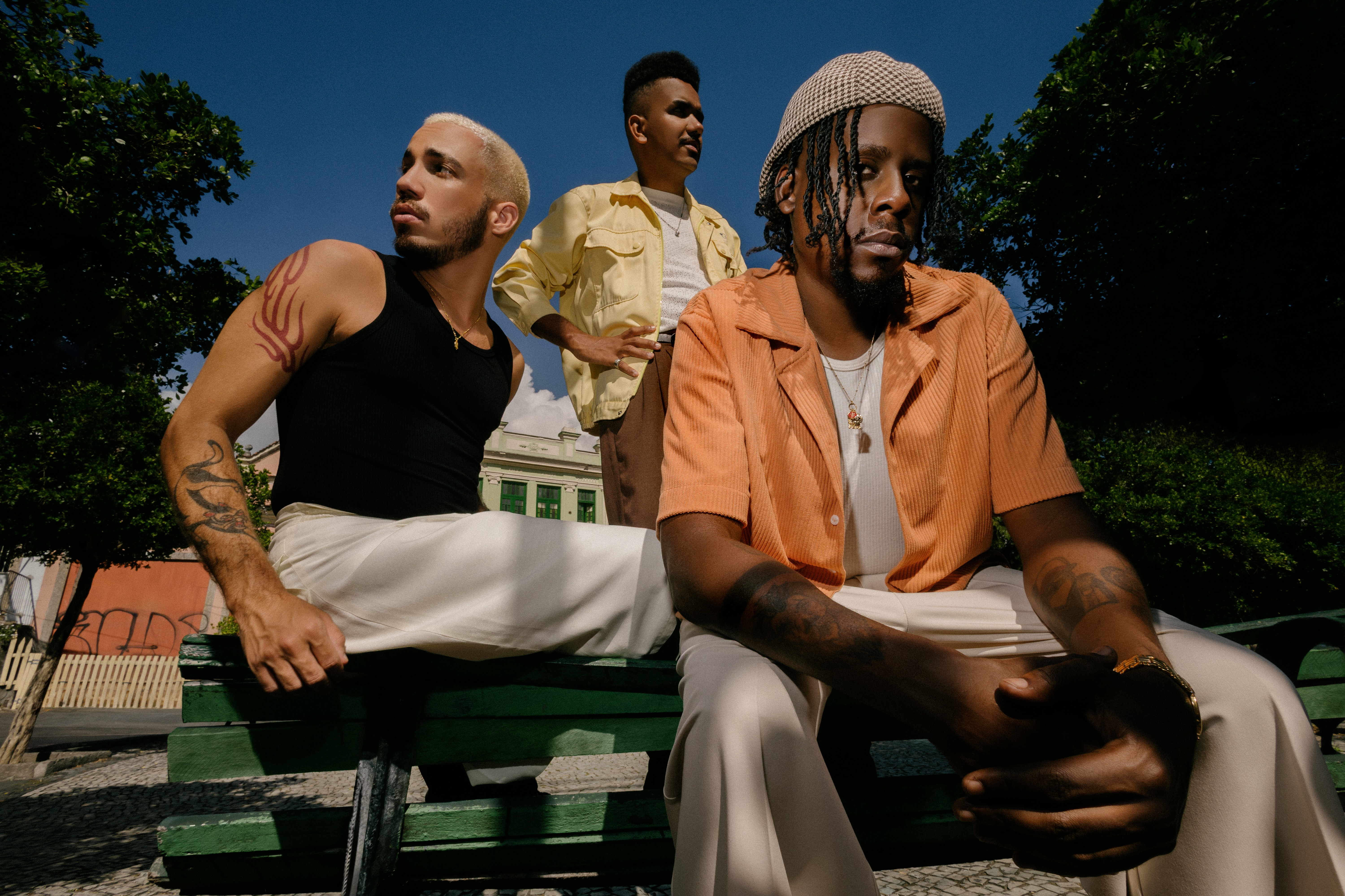 Três homens jovens, um branco e dois negros, posam em um banco verde ao ar livre. O homem branco, à esquerda, tem cabelo loiro platinado, veste regata preta e calça clara, com tatuagens nos braços. O homem negro do centro, em segundo plano, usa jaqueta amarela e calça marrom. O homem negro da direita, em primeiro plano, usa um chapéu de crochê, camisa laranja, camiseta branca e calça clara, com tatuagens nos braços e um colar. O céu azul e árvores verdes compõem o fundo