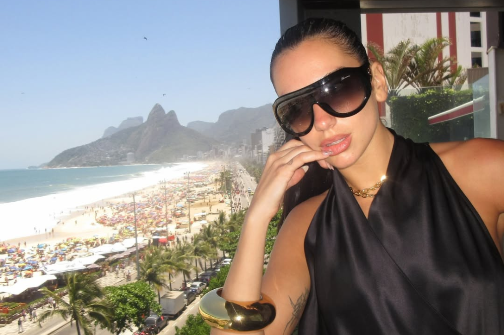 Mulher de óculos escuros grandes e blusa preta, com colar dourado e bracelete, posa em varanda com vista para a praia de Ipanema, Rio de Janeiro, cheia de guarda-sóis e pessoas, com o Morro Dois Irmãos ao fundo