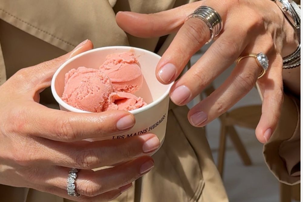 Mãos femininas segurando um copo branco com três bolas de sorvete rosa, unhas com esmalte nude e anéis variados nos dedos, incluindo um solitário e um de aliança com diamantes