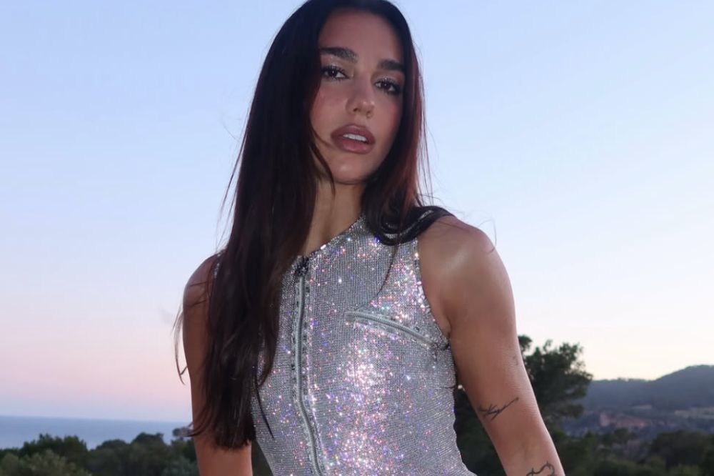 Dua Lipa, com cabelos longos e escuros, usa um top prateado brilhante com zíper frontal, olhando diretamente para a câmera. Ao fundo, um céu azul claro e vegetação escura, com o mar visível no horizonte