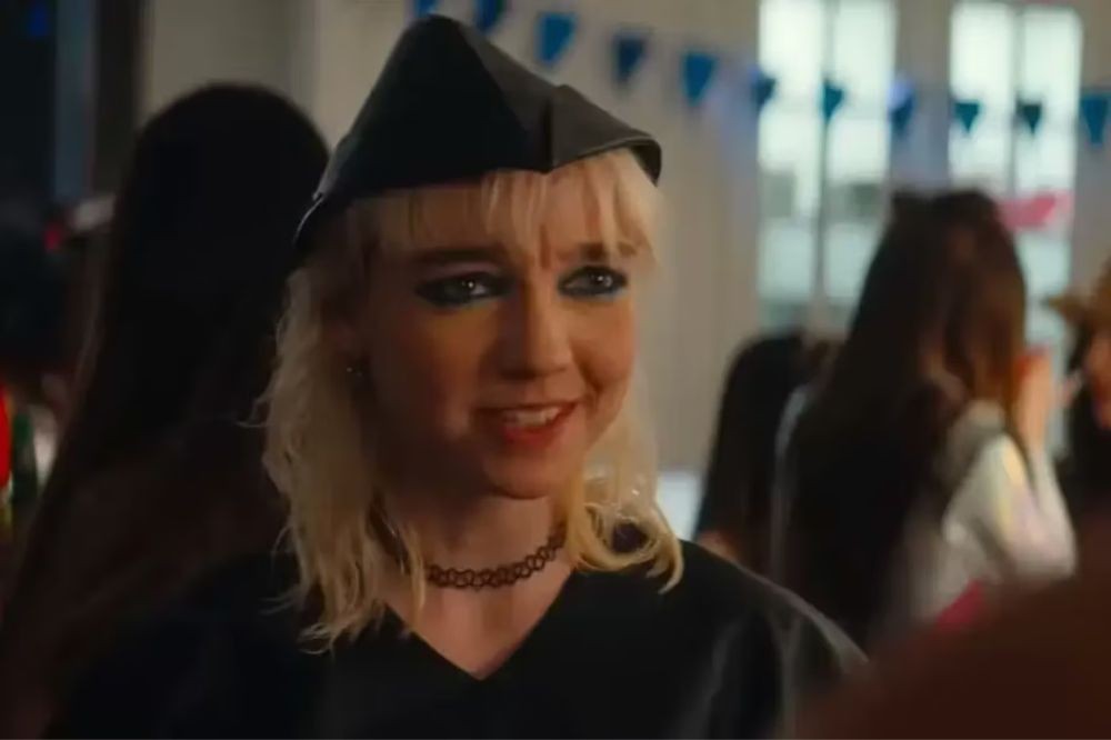 Jovem loira de pele clara, com chapéu preto pontudo, maquiagem escura nos olhos e gargantilha preta, sorrindo em uma festa com bandeirinhas azuis ao fundo