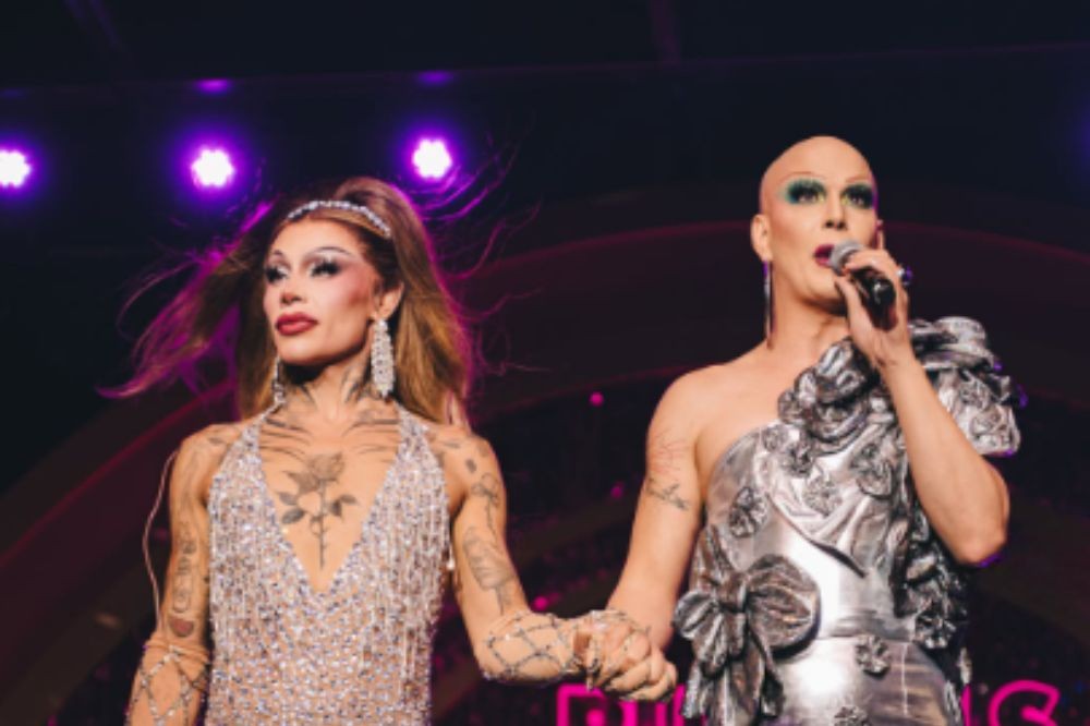 Duas drag queens no palco, uma com cabelo comprido e vestido prateado brilhante, a outra careca com maquiagem vibrante e vestido prateado com laços, segurando um microfone e cantando
