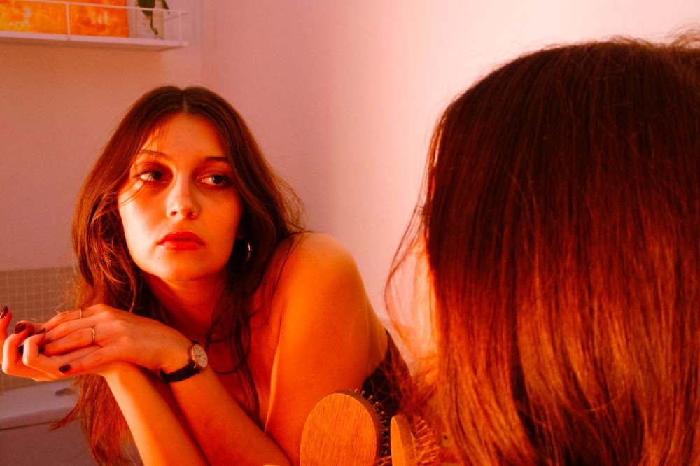 Mulher jovem de cabelos castanhos longos, com expressão pensativa, olhando para o lado esquerdo, refletida em um espelho. Ela usa um relógio de pulso e tem as mãos cruzadas. A iluminação avermelhada cria um ambiente íntimo e introspectivo