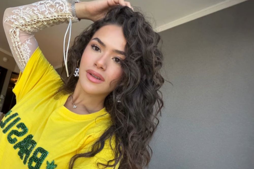 Mulher jovem de cabelos castanhos cacheados, com maquiagem leve e batom rosa, vestindo uma camiseta amarela com a palavra BRASIL em verde brilhante e uma manga de renda branca, posando com a mão direita nos cabelos