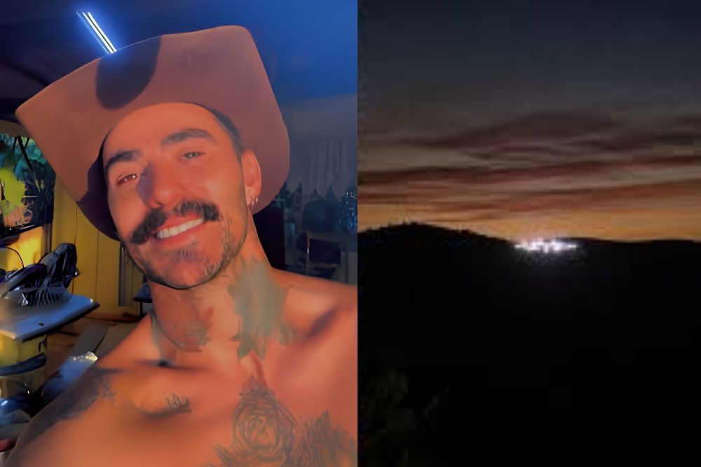 Homem sorridente de chapéu marrom e bigode, sem camisa, com tatuagens no pescoço e peito, ao lado de uma paisagem noturna com montanhas escuras e luzes distantes sob um céu alaranjado