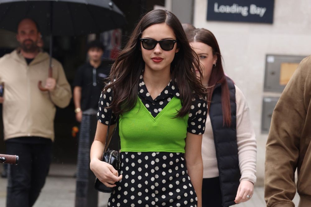 Mulher jovem de cabelos escuros e óculos de sol, vestindo um vestido preto de bolinhas brancas e um colete verde-limão, caminhando na rua com uma bolsa preta. Pessoas desfocadas ao fundo
