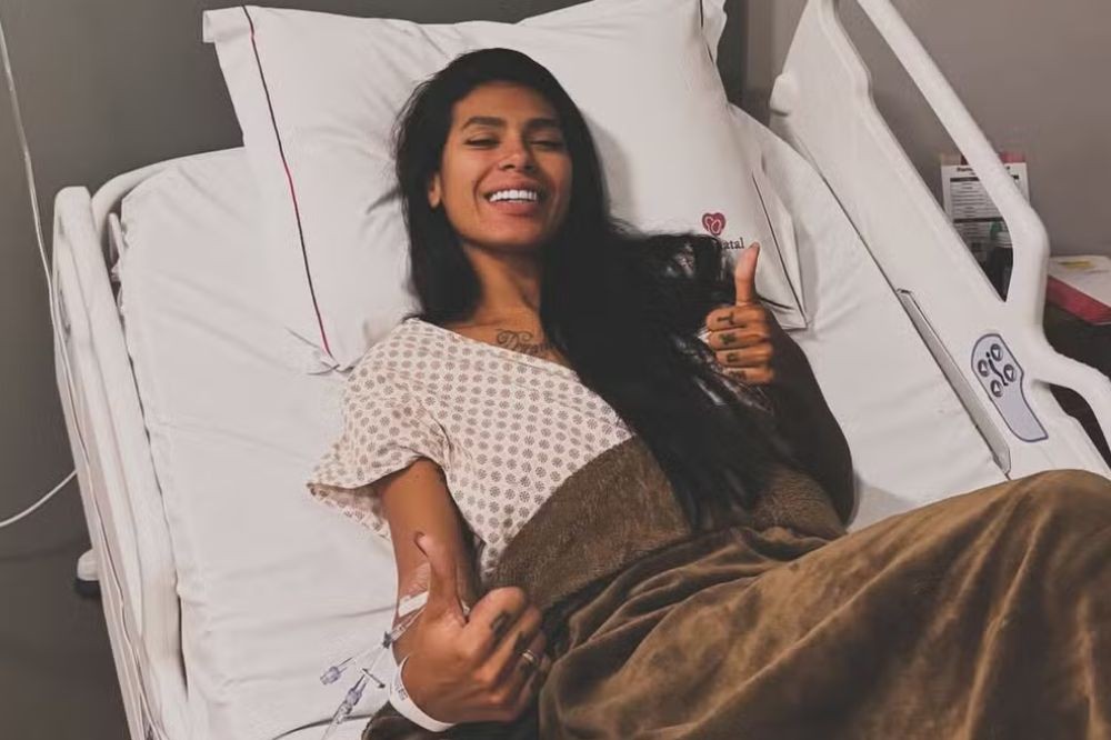 Mulher de cabelos longos e escuros, deitada em uma cama de hospital, sorrindo e fazendo sinal de positivo com as duas mãos. Ela veste uma camisola hospitalar e está coberta por um cobertor marrom. Um acesso venoso está visível em seu braço esquerdo.