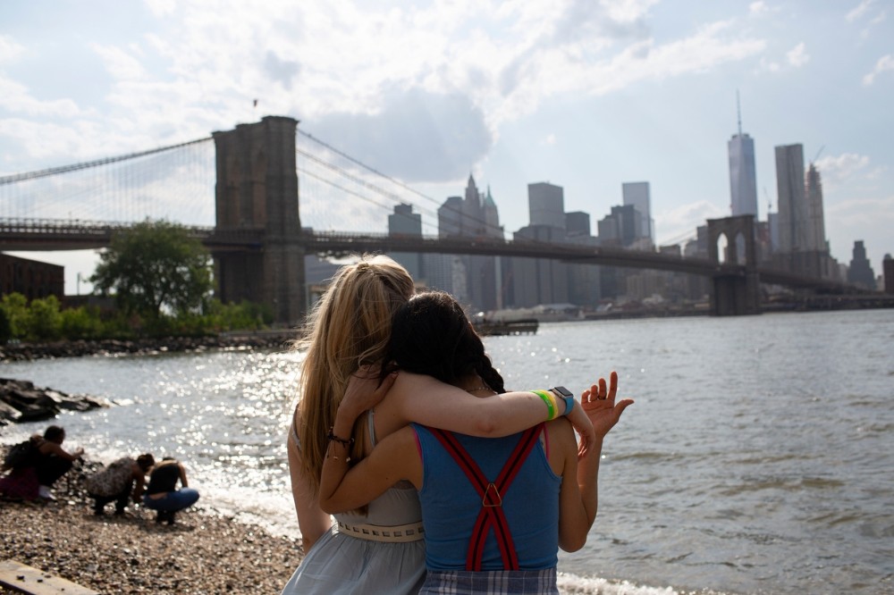 Duas mulheres abraçadas, de costas, observam o rio com o Brooklyn Bridge e o horizonte de Nova York ao fundo. O sol reflete na água, e o céu tem nuvens esparsas