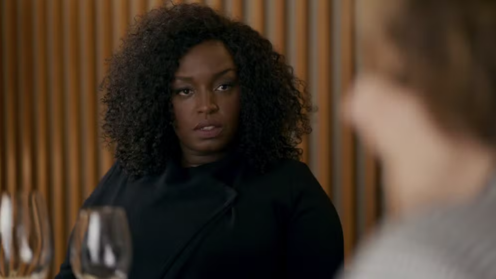 Mulher negra de cabelo cacheado escuro, vestindo casaco preto, olha com expressão séria para alguém fora do enquadramento, com taças de vinho em primeiro plano e parede de madeira ao fundo