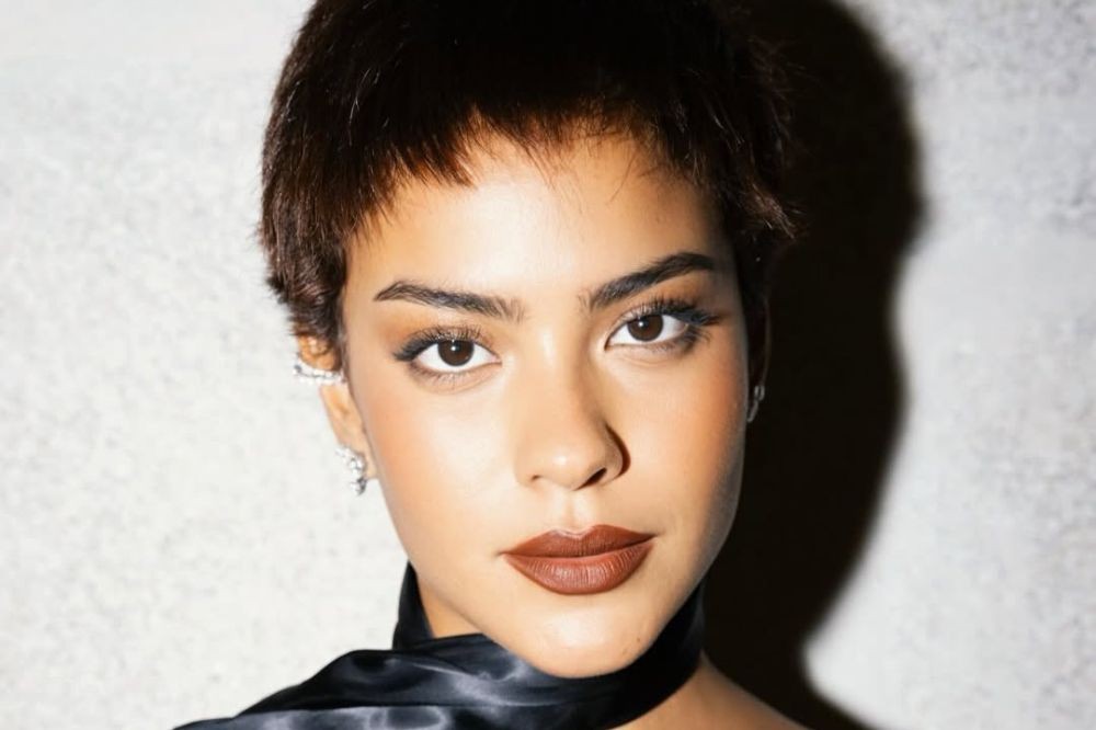Mulher jovem com cabelo castanho curto e franja, maquiagem leve, batom marrom, brincos e colar de tecido preto, olhando diretamente para frente