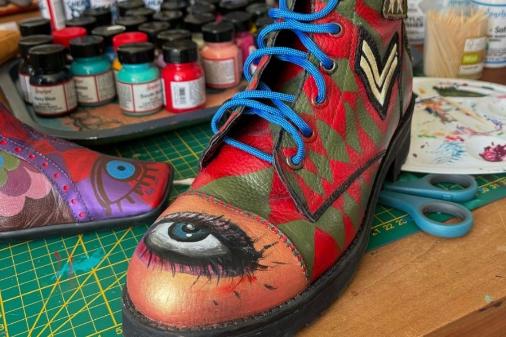 Bota de couro com cadarços azuis, pintada com um olho grande na ponta e padrões geométricos vermelhos e verdes no corpo, sobre uma base de corte verde com tesoura e tintas ao fundo