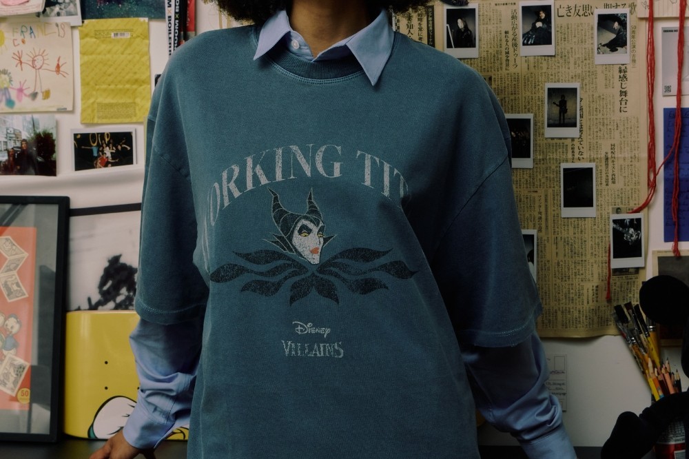 Pessoa com blusa de moletom azul-petróleo sobre camisa azul-clara, com estampa da Malévola e texto Disney Villains. Ao fundo, parede com desenhos infantis, fotos e recortes de jornal