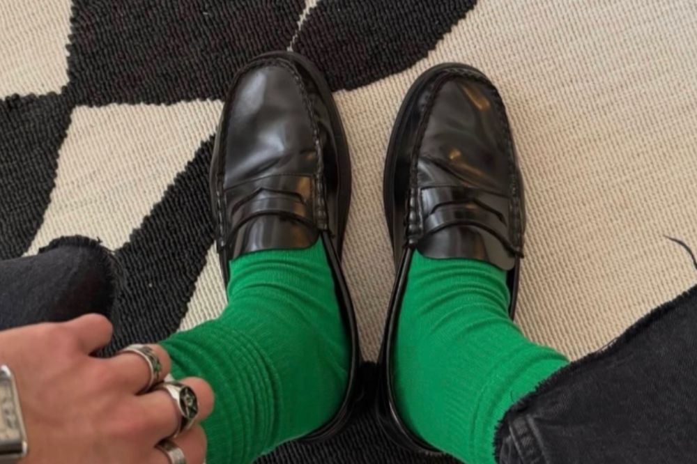 Pés de uma pessoa usando meias verdes vibrantes e sapatos mocassim pretos brilhantes, sobre um tapete com estampa geométrica preta e branca. A mão esquerda, com anéis prateados e um relógio, aparece no canto inferior esquerdo, e a barra de uma calça jeans preta rasgada no canto inferior direito