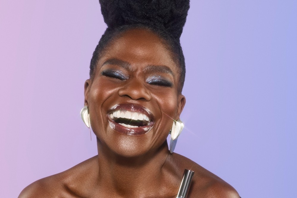 Mulher negra sorrindo amplamente com os olhos fechados, cabelo crespo preso no alto, maquiagem prateada nos olhos, gloss nos lábios e brincos prateados grandes, em fundo lilás e azul