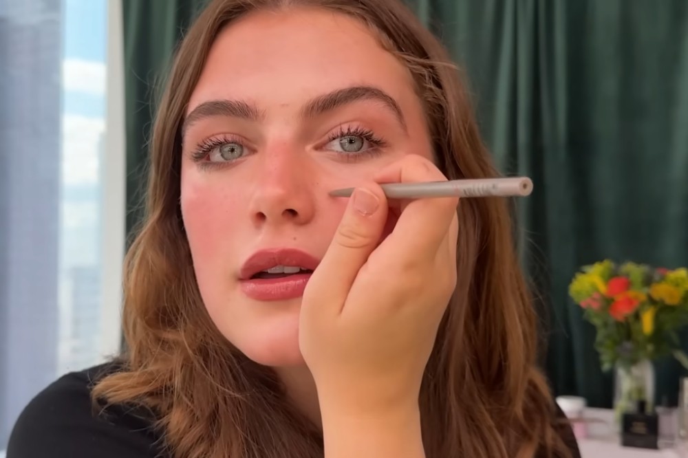 Mulher jovem, de cabelos castanhos e olhos azuis, aplicando maquiagem no rosto. Ela segura um lápis cinza claro próximo ao canto interno do olho esquerdo, com a boca entreaberta. O fundo mostra uma cortina verde escura e um arranjo de flores coloridas