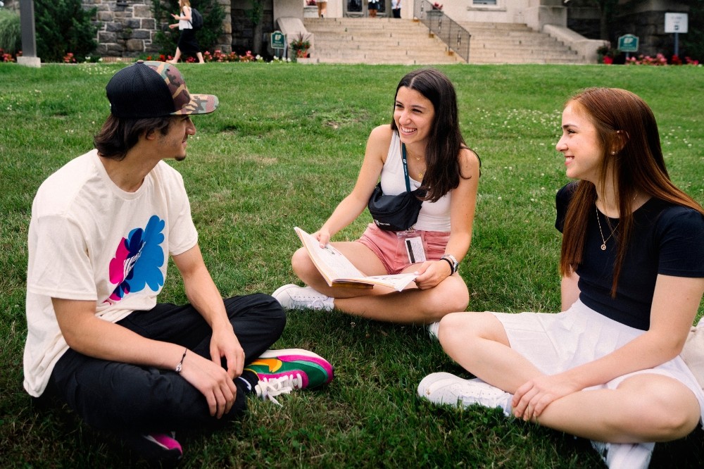 Três jovens sentados na grama conversando. Um rapaz de boné e camiseta clara com estampa colorida, uma moça de regata branca e shorts rosa segurando um livro, e outra moça de camiseta preta e saia branca, todos sorrindo