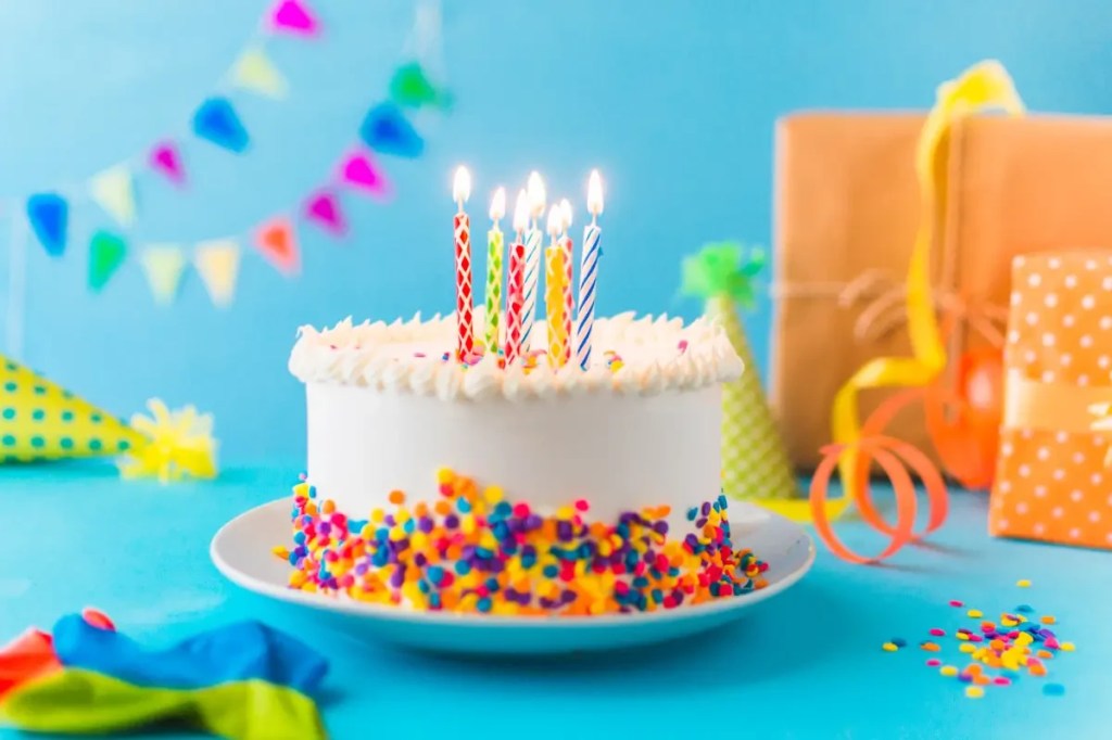Bolo de aniversário branco com cobertura de glacê e granulado colorido, com seis velas acesas. Ao fundo, presentes embrulhados, balões e chapéus de festa, sobre uma mesa azul