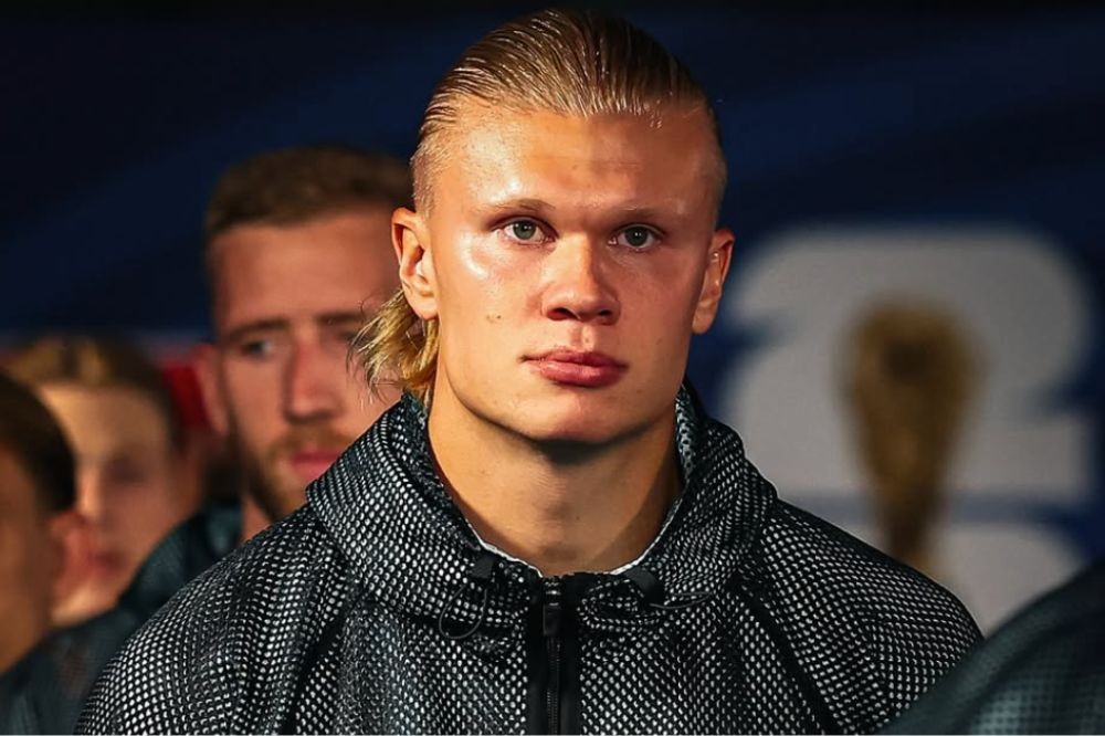 O jogador de futebol Erling Haaland, com cabelo loiro penteado para trás e mullet, veste uma jaqueta preta de tecido furado, olhando sério para frente. Outras pessoas desfocadas aparecem ao fundo