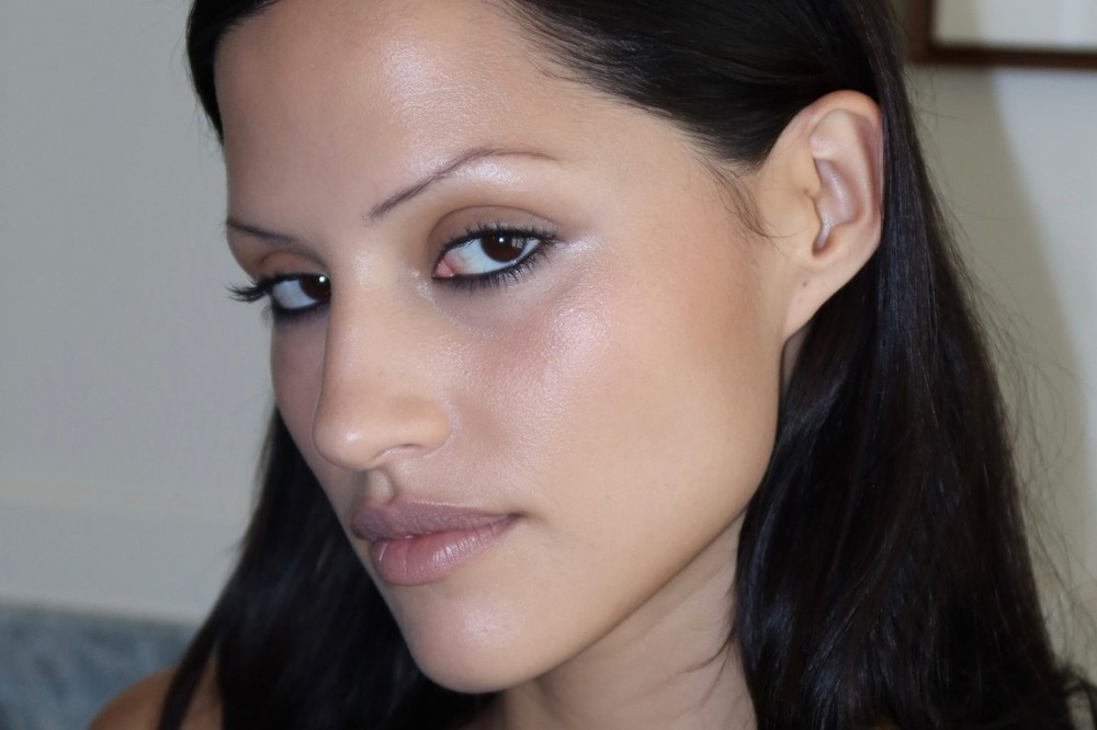 Close-up de uma mulher com pele morena clara e cabelos escuros, olhando para a câmera com maquiagem nos olhos e lábios. Ela tem delineador preto e cílios longos, com lábios em tom nude.