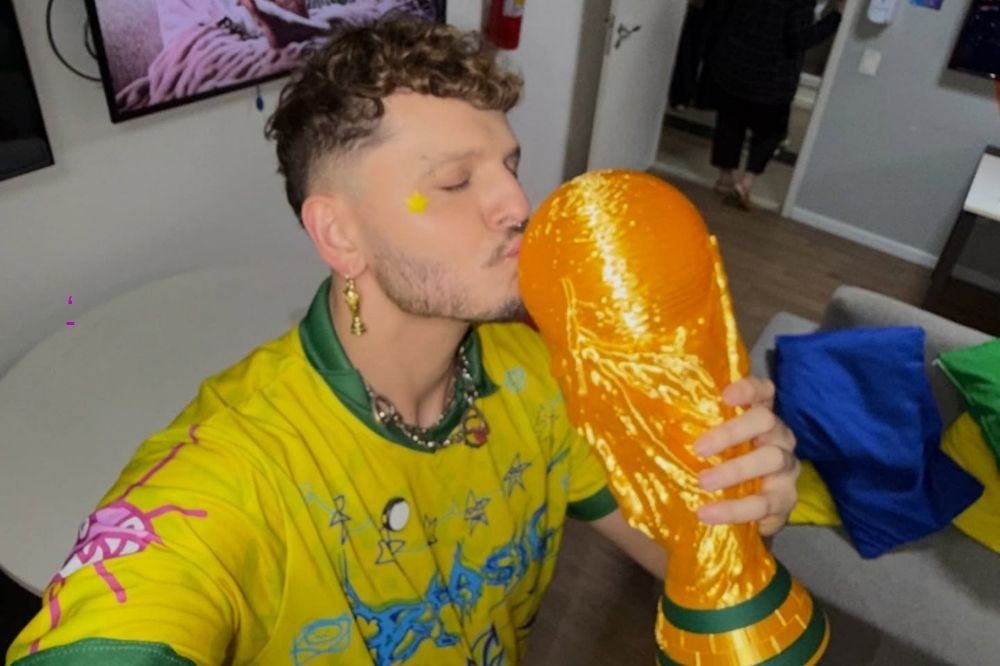 Homem jovem de cabelo cacheado e barba rala, usando camisa amarela da seleção brasileira e brinco dourado, beija uma réplica da taça da Copa do Mundo com os olhos fechados