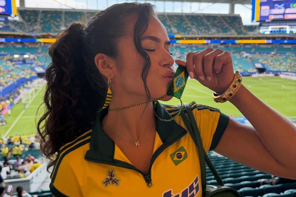 Mulher de cabelo escuro preso, vestindo camisa amarela e verde do Brasil, beijando um chaveiro com a bandeira brasileira, em um estádio de futebol lotado