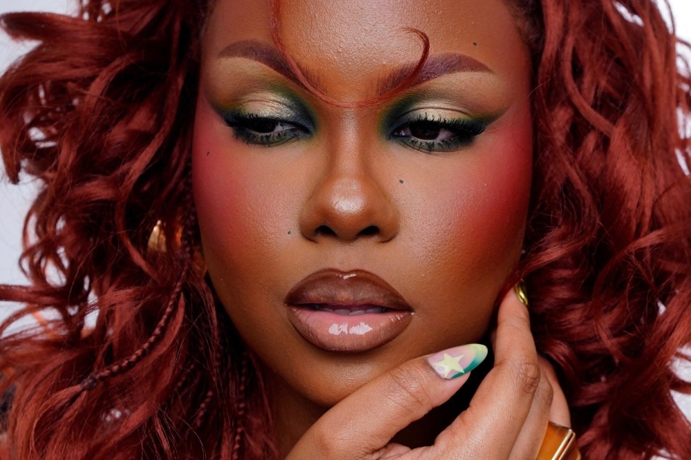 Mulher negra com cabelo ruivo cacheado, maquiagem vibrante com sombra verde e dourada, blush rosa intenso e lábios glossy. Ela toca o queixo com a mão, exibindo unhas decoradas com estrelas coloridas