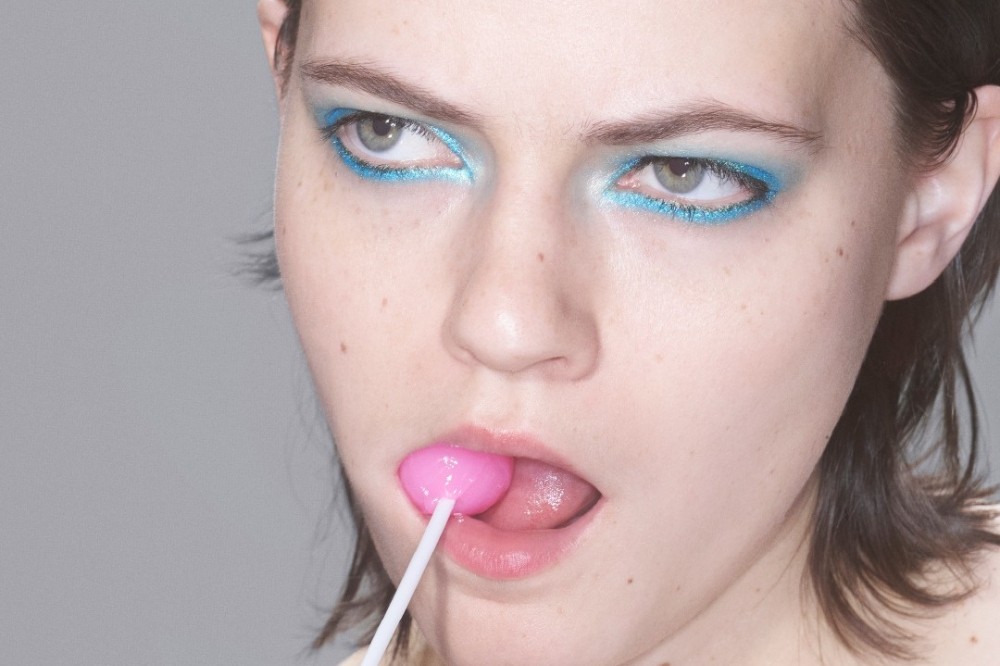 Rosto de uma pessoa com maquiagem azul brilhante nos olhos, lambendo um pirulito rosa com a língua para fora, olhando para cima e para a direita