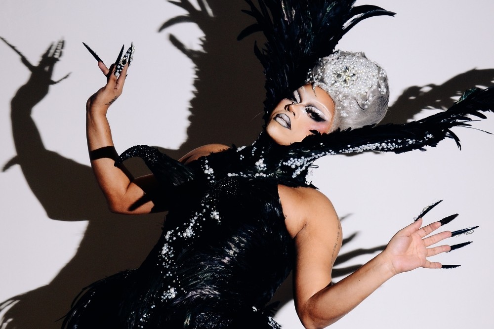 Artista drag com maquiagem dramática, peruca prateada e adereço de cabeça com pérolas, vestindo um traje preto de penas e pérolas, com unhas postiças longas e pretas, posando com os braços levantados contra uma parede branca com sombras marcantes