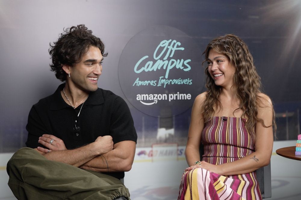 Um homem e uma mulher sorrindo, sentados um de frente para o outro. Ele tem cabelo cacheado escuro, usa camisa polo preta e calça verde-oliva. Ela tem cabelo ondulado castanho, veste um vestido tomara que caia listrado em tons de vinho, rosa e amarelo. Ao fundo, um banner com o logo Off Campus Amores Improváveis Amazon Prime
