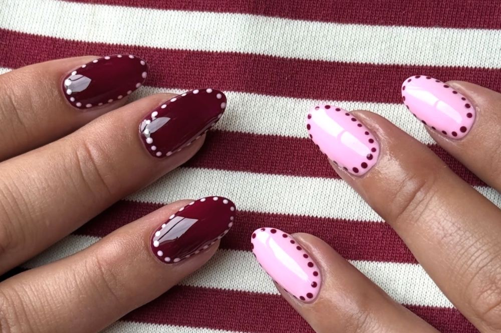Mãos com unhas decoradas: três unhas em tom vinho com bolinhas brancas na borda e duas unhas em rosa claro com bolinhas vinho na borda, sobre tecido listrado branco e vinho