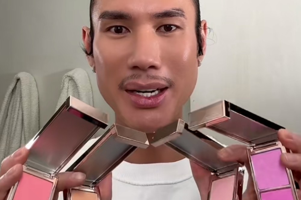 Homem asiático com bigode e cavanhaque, maquiado, segurando cinco paletas de maquiagem abertas com blushes e iluminadores em tons de rosa, pêssego e lilás, contra um fundo claro com toalhas brancas
