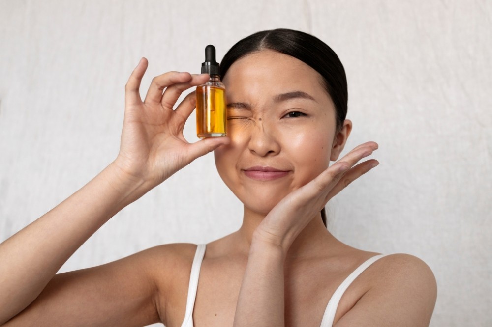 Mulher asiática sorrindo, com cabelo preso, segurando um frasco de conta-gotas com líquido dourado sobre o olho esquerdo, que está fechado e enrugado. A mão direita está na bochecha, e ela veste uma alça branca de sutiã, em fundo branco