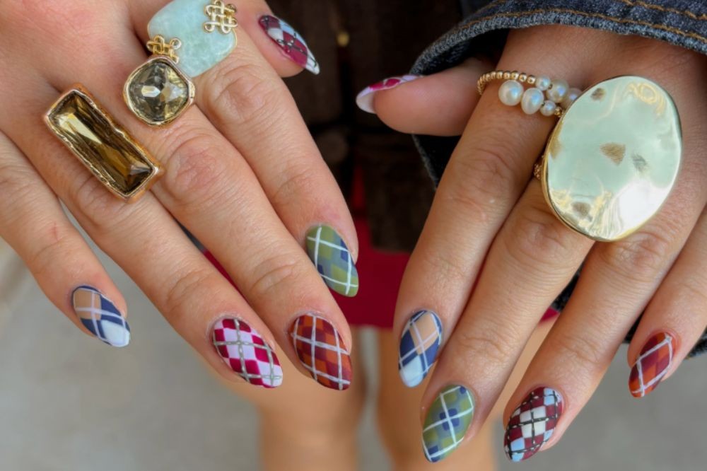 Mãos femininas com unhas longas e pontudas, decoradas com padrões xadrez em tons de azul, verde, marrom e vinho. Vários anéis dourados com pedras grandes adornam os dedos