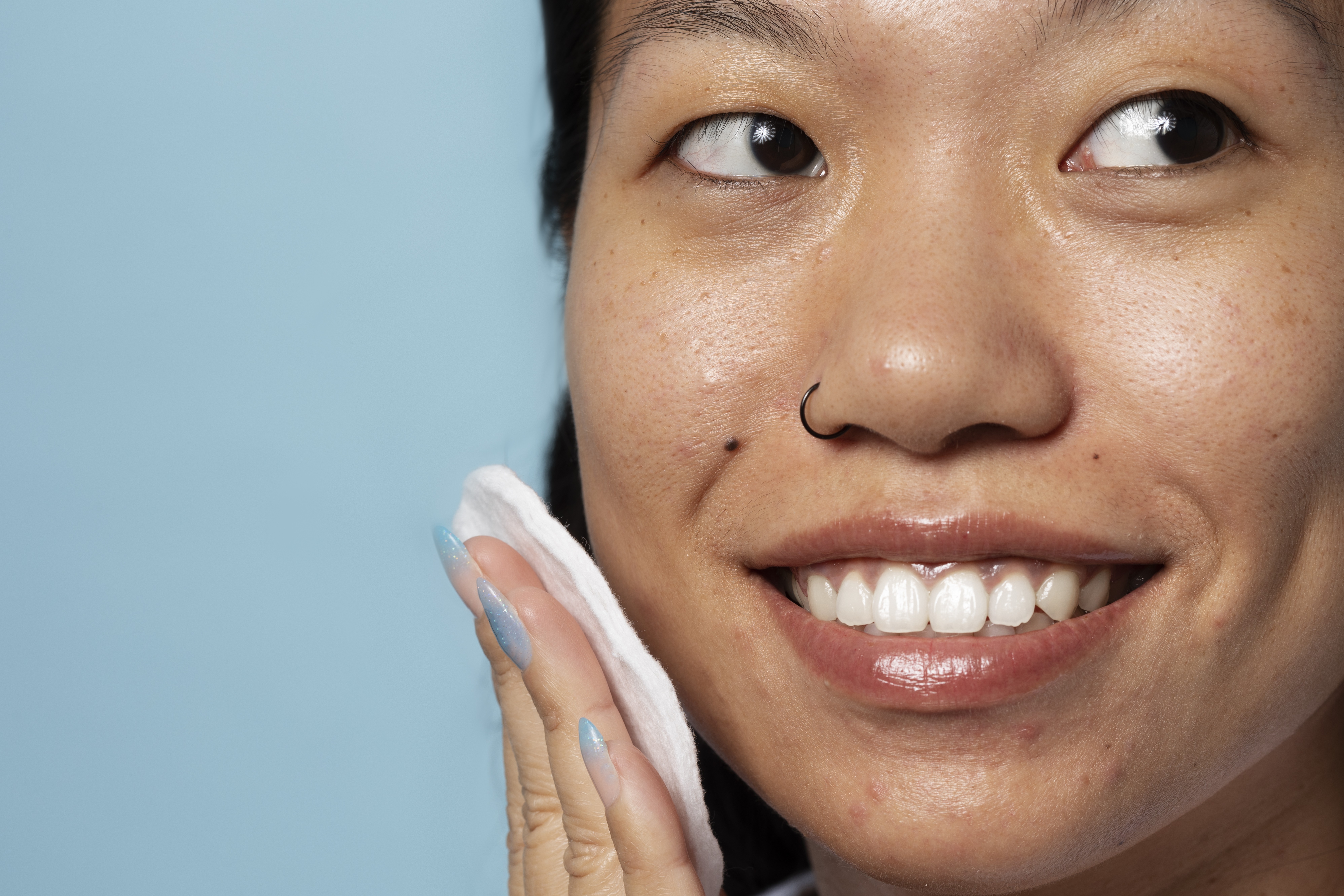 Mulher asiática sorrindo, com piercing no nariz, aplicando produto no rosto com algodão. Ela tem unhas longas e azuis, e a pele com algumas marcas e poros visíveis. O fundo é azul claro
