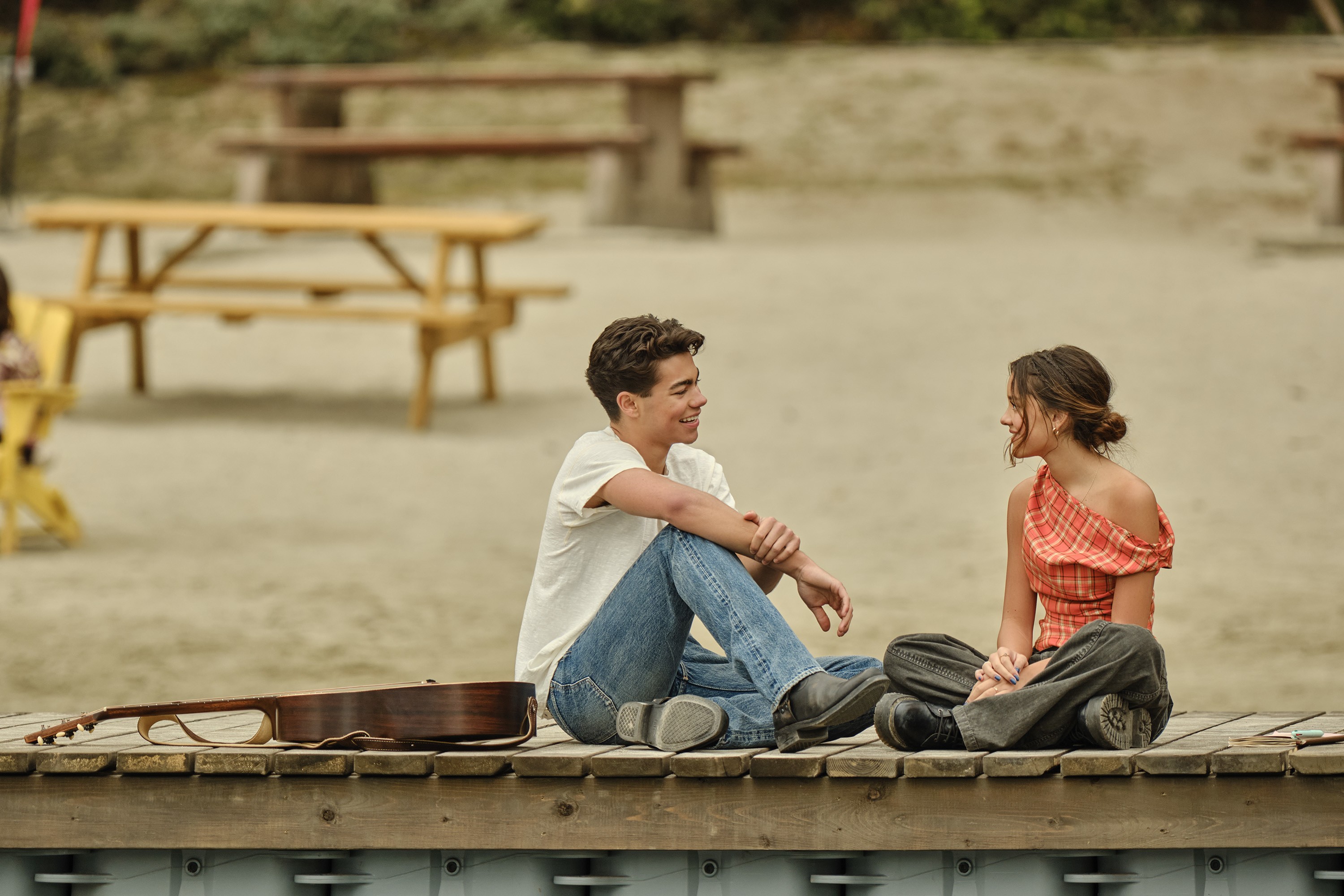 Um jovem casal sorridente sentado em um píer de madeira, conversando. O rapaz veste camiseta branca e jeans, a moça usa blusa xadrez laranja e calça escura. Um violão marrom está deitado no píer, e ao fundo, mesas de piquenique em uma área de areia.