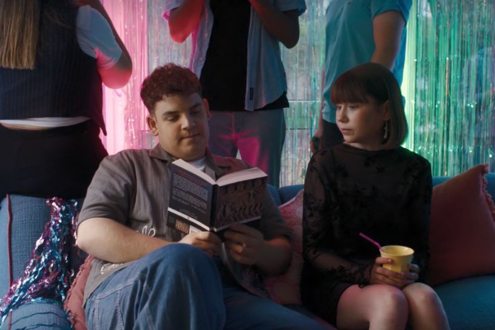 Um homem jovem com cabelo encaracolado e camisa cinza lê um livro enquanto uma mulher de cabelo curto e franja, usando vestido preto, segura um copo amarelo com canudo rosa, ambos sentados em um sofá azul em uma festa com decoração brilhante