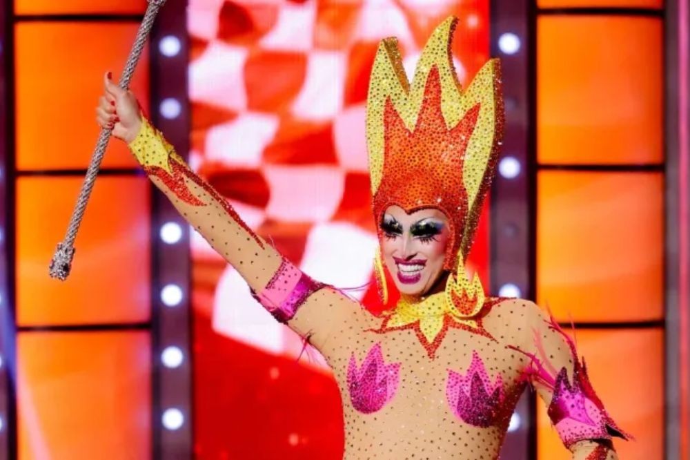 Drag queen sorridente, com maquiagem marcante e batom roxo, usa um body nude cravejado de brilhos, com detalhes em rosa e amarelo. Ela veste uma coroa e brincos em formato de chamas, nas cores laranja e amarelo, e segura um bastão prateado com a mão direita erguida. O fundo é um palco com painéis quadriculados em vermelho e branco, e outros em laranja