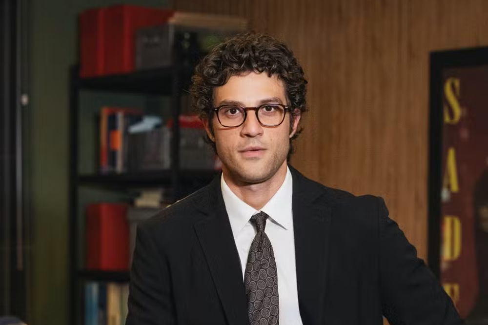 Homem jovem de pele clara, cabelo cacheado escuro e óculos, vestindo terno preto, camisa branca e gravata estampada, olhando para frente com expressão séria. Ao fundo, uma estante com livros e objetos, e uma parede de madeira