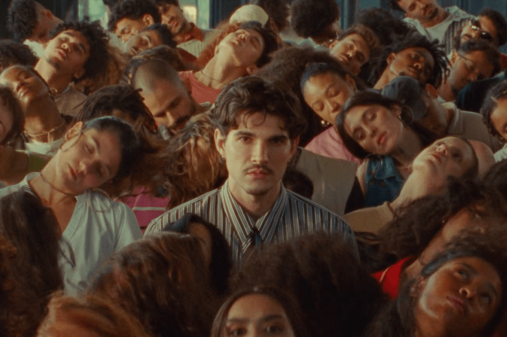 Um homem com bigode e camisa listrada olha fixamente para frente, cercado por diversas pessoas deitadas, com os olhos fechados e cabeças inclinadas para trás ou para os lados, em um ambiente com luz suave