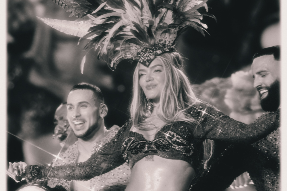 Cantora loira com cocar de penas e top brilhante, sorrindo e dançando entre dois homens sorridentes, em preto e branco