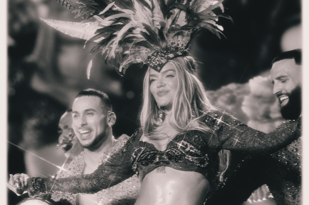 Cantora loira com cocar de penas e top brilhante, sorrindo e dançando entre dois homens sorridentes, em preto e branco